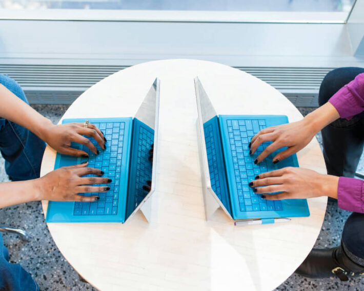 Two sets of hands facing each other typing on laptop keyboards sitting at a shared table.