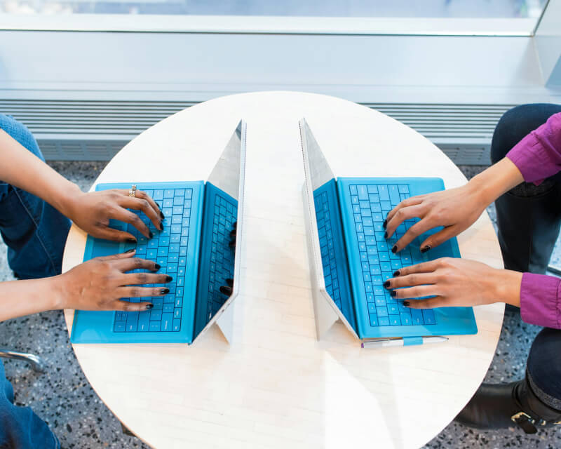 Two sets of hands facing each other typing on laptop keyboards sitting at a shared table.