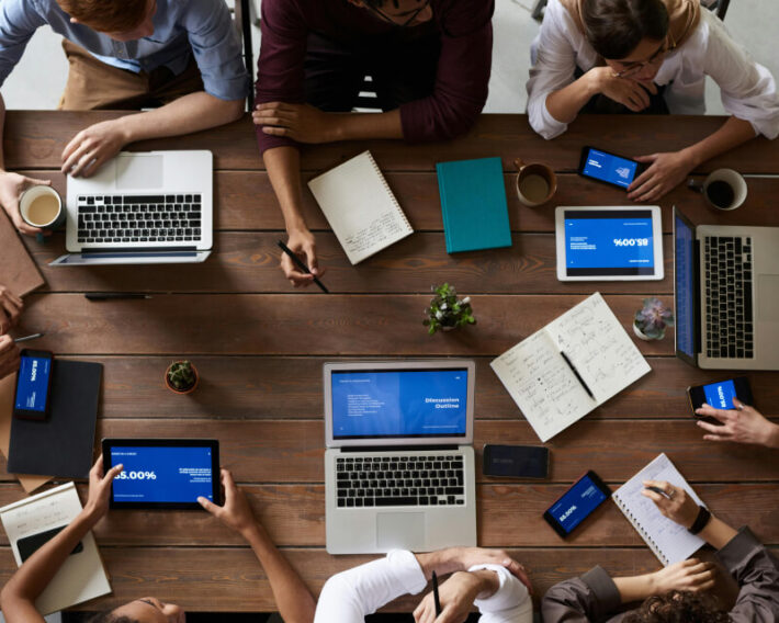 Eight people with laptops collaborating around at a wood desk.
