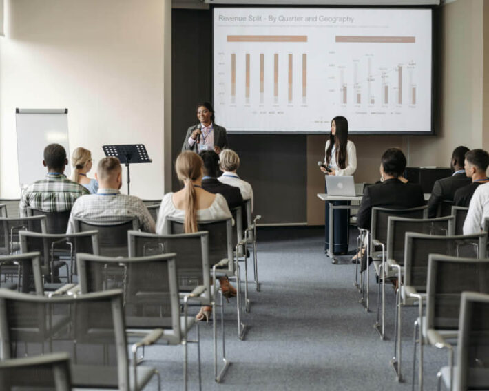 Person standing referencing a presentation on a screen while three seated people listen.