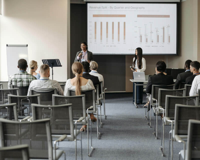 Person standing referencing a presentation on a screen while three seated people listen.